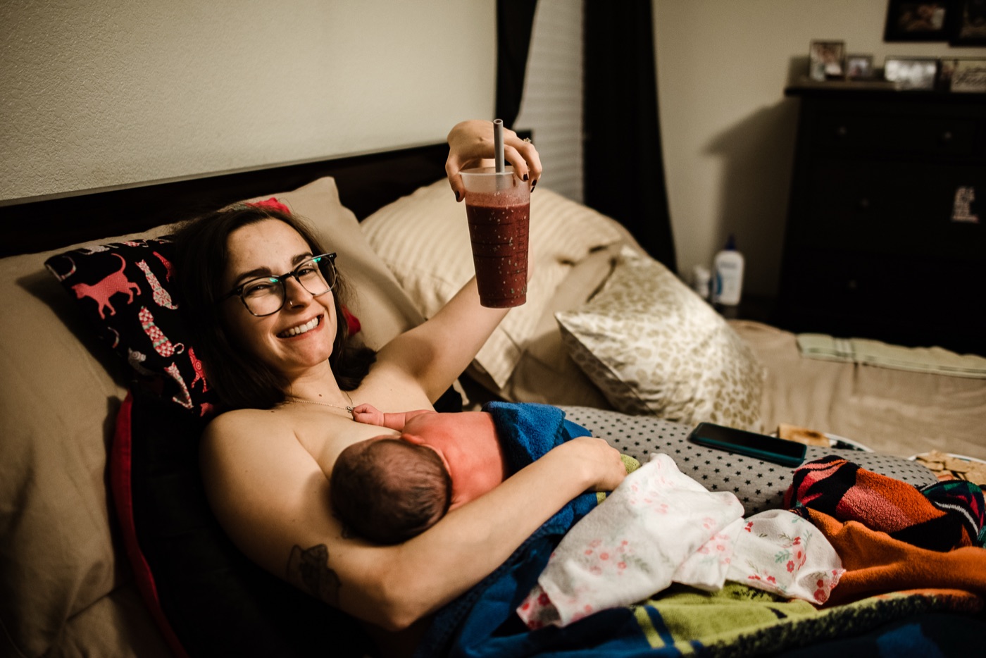 A new mother resting in bed, nursing her newborn and holding a smoothie